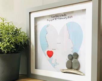 Op de afbeelding: Een ingelijste hartvormige kaart met een rood hart dat een locatie markeert. De kaart is omgeven door een grijs frame en bevat de tekst "Lans Eric & Kelly Verloofd op 10 februari 2021".
