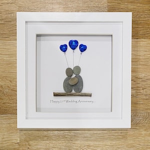Op de afbeelding: Een ingelijst kunstwerk van kiezelsteen dat een stel toont dat elkaar vasthoudt met blauwe hartballonnen erboven. De tekst "Happy 21st Wedding Anniversary..." staat onder het stel.