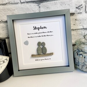 Puede incluir: Una pieza de arte de guijarros enmarcada con un marco gris y un fondo blanco. El arte presenta dos guijarros grises que representan personas sentadas en un pequeño trozo de madera a la deriva. El texto dice "Stephen There's no better friend than a Brother, And there's no better Brother than you... With love from Karen XX".