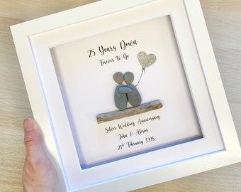 May include: A framed pebble art piece celebrating a 25th wedding anniversary. The art features two figures holding hands, a heart-shaped balloon, and the text "25 Years Down Forever to Go". The text "Silver Wedding Anniversary John & Alison 21st February 1998" is also included.