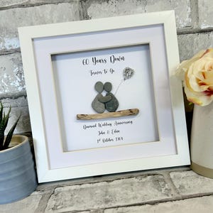 Op de afbeelding: Een wit ingelijst beeld met een kiezelsteenkunstontwerp van een stel dat elkaars hand vasthoudt. De tekst luidt "60 Years Down Forever to Go Diamond Wedding Anniversary John & Edna 1st October 1964".
