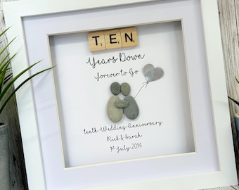 May include: A white framed picture with a white background. The picture features a couple made of gray stones holding a silver heart balloon. The text "T. E, N Years Down forever to Go" is written in black cursive. The text "tenth Wedding Anniversary Rick & Sarah 21 July 2014" is written in black cursive.