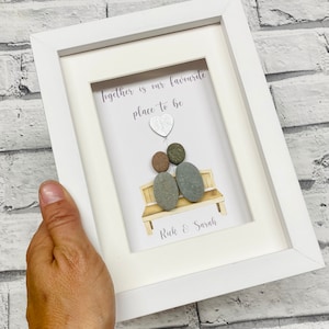 Op de afbeelding: Witte ingelijste afbeelding met een kiezelsteenkunstontwerp van twee mensen die op een bankje zitten. De tekst luidt "Together is our favorite place to be" met een hartballon boven het paar. De namen "Rick & Sarah" staan onder het paar.