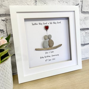 Può includere: Un'opera d'arte incorniciata con una coppia fatta di pietre, che tiene un palloncino rosso a forma di cuore. Il testo recita "Together they lived a life they loved. John & Katie Ruby Wedding Anniversary. 10th June 1983."
