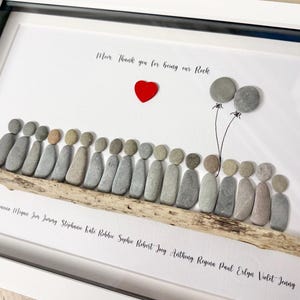 Puede incluir: Obra de arte enmarcada con fondo blanco, con una base de madera flotante con figuras de guijarros. Un corazón rojo y globos de guijarros están encima de las figuras. El texto dice "Mamá, gracias por ser nuestra roca" y nombres debajo.