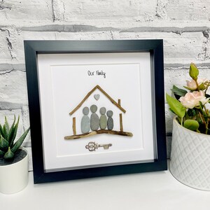 May include: A framed pebble art piece depicting a house with a heart above it and five figures inside. The text "Our Family" is written above the house. A silver key is below the house.