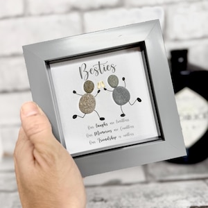 Puede incluir: Una obra de arte enmarcada con un borde gris, con la palabra "Besties" y dos figuras de guijarros sosteniendo copas de champán. El texto de abajo dice "Nuestras risas son incontables, Nuestros recuerdos son incontables, Nuestra amistad es interminable."
