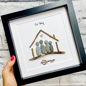 May include: A framed pebble art piece depicting a house with a family of five inside. The house is made of driftwood and the family members are made of gray pebbles. The words "Our Family" are written in black ink at the top of the frame.