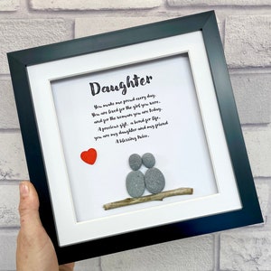 May include: A framed artwork with the word "Daughter" in script. The artwork features a red heart and two figures made of gray stones on a branch. The text reads, "You make me proud every day..." in black ink.