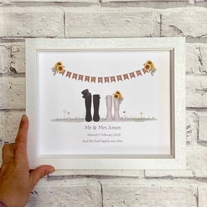 Puede incluir: Una impresión enmarcada en blanco con una ilustración de boda de dos pares de botas de lluvia, una negra y una blanca, con una pancarta que dice "Just Married". El texto debajo dice "Mr. & Mrs. Jones Casados el 1 de febrero de 2023 Y vivieron felices para siempre..."