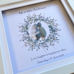 May include: A framed pebble art piece depicting a couple inside a floral wreath. The text reads "Mr & Mrs Anderson", "Love, laughter & happily ever after.." and "Saturday 17th June 2023". The frame is white.