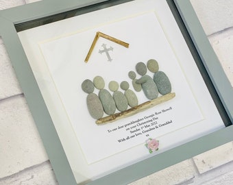 Könnte beinhalten: Ein gerahmtes Bild aus Kieselsteinen mit einem Kreuz und einem Holzdach, das eine Kirche darstellt. Die Kieselsteine sind so angeordnet, dass sie eine Familie von Menschen bilden, die auf einem Treibholzast sitzen. Der Text lautet "To our dear granddaughter Georgie Rose Showell on your Christening Day Sunday 1st May 2022 With all our love, Grandma & Granddad xx".