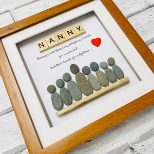 Puede incluir: Una obra de arte enmarcada con la palabra "NANNY" deletreada con fichas de letras de madera. La obra de arte también incluye una representación de una familia en guijarros con un corazón rojo. El texto dice "Nanny's hold their Grandchildren's hands for a moment and their hearts for a lifetime..."