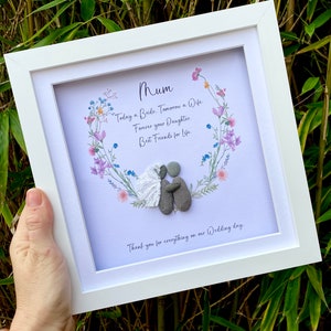 Puede incluir: Una tarjeta blanca enmarcada con una corona floral de acuarela y una pareja de arte de guijarros en el interior. La tarjeta dice "Mamá, Hoy una novia. Mañana una esposa. Para siempre tu hija. Mejores amigas para la vida. Gracias por todo en nuestro día de boda."
