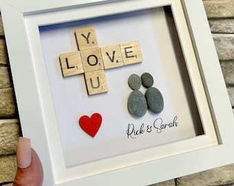 May include: A white framed picture with a red heart, two gray pebble figures, and a wooden tile crossword puzzle spelling "I LOVE YOU". The picture is signed "Rick & Sarah".