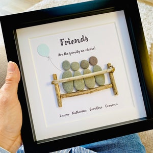 Op de afbeelding: Een ingelijst kunstwerk met een kiezelsteenkunstontwerp van vier vrienden die op een bankje zitten. Het kunstwerk heeft de tekst "Friends are the family we choose!" en de namen "Laura, Katherine, Caroline, Gemma" staan onder het bankje.