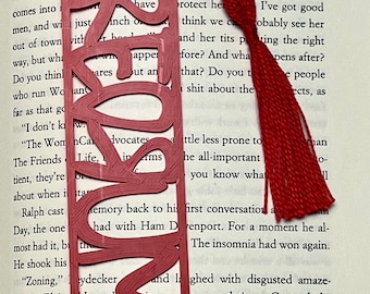 Puede incluir: Un marcapáginas rojo con un diseño decorativo y un borla. El marcapáginas está hecho de plástico y tiene un diseño recortado. El marcapáginas está colocado en un libro con texto.