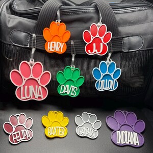 May include: A selection of brightly coloured paw-print pet tags, each with a name such as "Henry" or "Luna." The tags are in shades of orange, red, pink, green, blue, burgundy, yellow, grey, and purple. Each tag has a clear plastic loop for attachment.