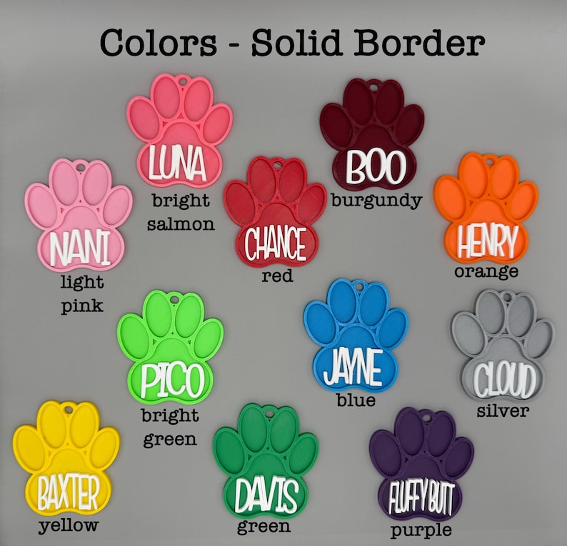 May include: A selection of brightly coloured paw-shaped pet tags. The tags are in shades such as light pink, bright salmon, red, burgundy, orange, blue, silver, yellow, green, and purple, each with a name printed on it.