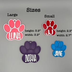May include: A selection of pet ID tags shaped like paw prints. The tags are in pink, red, purple, and blue. The tags are labelled with names: LUNA, JJ, INDIANA, and JAYNE. The large tag is 9.9 cm wide and high, and the small tag is 6.9 cm wide and 7.4 cm high.