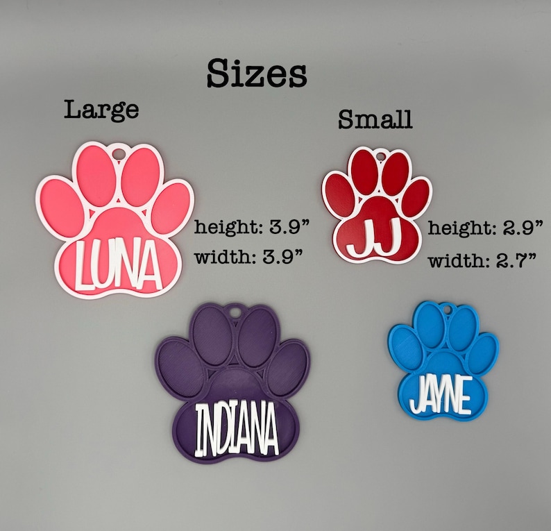 May include: A selection of pet ID tags shaped like paw prints. The tags are in pink, red, purple, and blue. The tags are labelled with names: LUNA, JJ, INDIANA, and JAYNE. The large tag is 9.9 cm wide and high, and the small tag is 6.9 cm wide and 7.4 cm high.