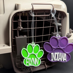 May include: Two paw print pet tags, one green and one purple, hang from a pet carrier. The green tag says "FLYNN" in white, and the purple tag says "INDIANA" in white. The tags are made of plastic.