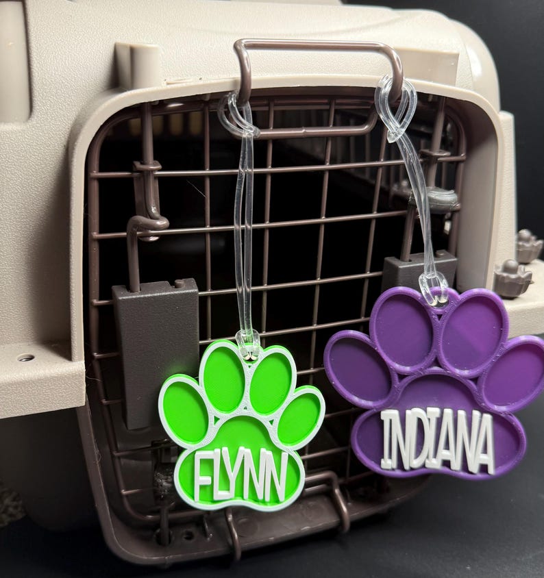 May include: Two paw print pet tags, one green and one purple, hang from a pet carrier. The green tag says "FLYNN" in white, and the purple tag says "INDIANA" in white. The tags are made of plastic.