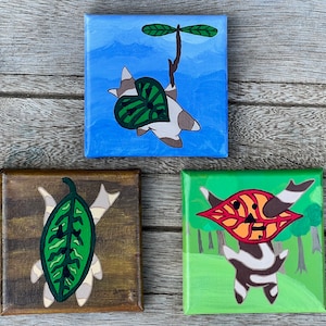 May include: Three small canvas paintings depicting stylised creatures with leaf-like features. The first painting is blue with a white creature with a green leaf on its back. The second painting is brown with a white creature with a green leaf on its back. The third painting is green with a white creature with a red leaf on its back.