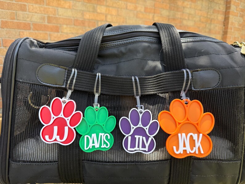 May include: A black pet carrier with four colourful paw-shaped tags. The tags are red, green, purple, and orange, each with a white name: JJ, DAVIS, LILY, and JACK. The tags are attached to the carrier.