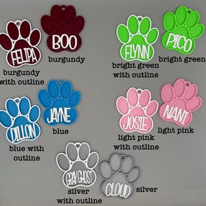 May include: A selection of paw-shaped pet tags in shades of burgundy, blue, green, pink, and silver. Each tag features a name, such as "FELIPA", "BOO", "FLYNN", "PICO", "DILLON", "JAYNE", "JOSIE", "NANI", "GRAY GHOST", and "CLOUD", with a contrasting outline.