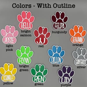 May include: A selection of colourful paw-shaped pet tags, each with a name and colour. The tags feature names such as "Bella" (bright salmon), "Josie" (light pink), and "Scrambles" (yellow). Each tag has a white outline and a hole for attachment.