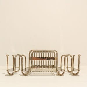 May include: A polished silver-tone metal toast rack featuring a central rectangular compartment with vertical wire dividers and two symmetrical, curved side sections designed to hold additional slices of toast in an upright position.