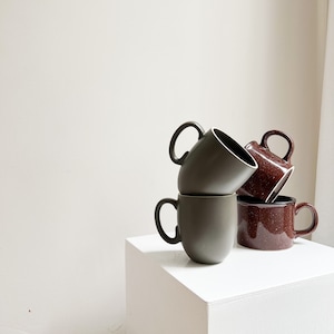 Könnte beinhalten: Ein Stapel aus drei Keramikbechern. Die beiden oberen Becher sind in einem gedämpften Grünton gehalten, während der untere Becher eine gesprenkelte braune Farbe hat.