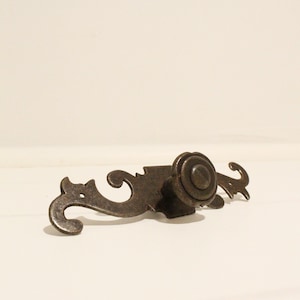 May include: A dark bronze-toned metal drawer pull featuring a central circular knob and an elongated, decorative backplate with curved, scroll-like ends. The hardware has a weathered, textured finish and two visible screw holes for mounting.