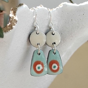 Può includere: Un paio di orecchini pendenti con ganci in argento, un piccolo elemento superiore circolare color crema e un pendente inferiore trapezoidale azzurro con un cerchio arancione centrato e un piccolo punto verde acqua al centro di ogni orecchino.