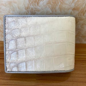 以下が含まれることがあります： 白のクロコダイルレザーで作られた二つ折り財布。財布は角が丸く、灰色の縁取りのある長方形です。ステッチは黒です。財布は木製の表面にあります。