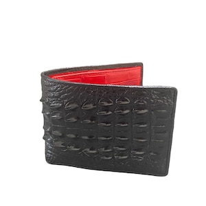 Puede incluir: Cartera negra de dos pliegues con textura de piel de cocodrilo. El interior es de un rojo vibrante. La cartera está abierta, mostrando el forro interior. El exterior tiene un acabado brillante.