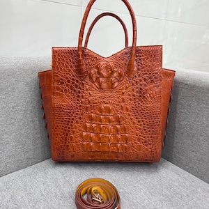 May include: A structured tote bag in a rich, burnt orange hue featuring a prominent embossed crocodile skin texture. The bag includes two top handles and a detachable, coiled shoulder strap with gold-toned metal hardware clips at each end.