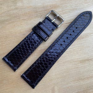 May include: A black watch strap with a textured, reptile-like pattern. The strap has a silver-colored buckle and is made of a shiny, dark material. The strap is shown in two pieces, ready to be attached to a watch.
