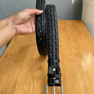 May include: Black belt with a crocodile-effect texture. The belt features a silver rectangular buckle and a matching loop. The belt is shown on a wooden surface.
