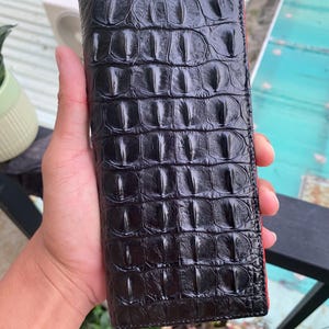 Puede incluir: Cartera negra con textura de piel de cocodrilo. La cartera rectangular tiene un interior rojo. La cartera está sujeta en la mano de una persona.