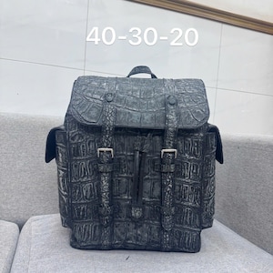 May include: A black backpack with a crocodile skin texture. The backpack has a flap closure with two buckle straps and a top handle. The dimensions are approximately 15.7 x 11.8 x 7.9 inches.