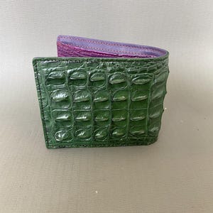 Puede incluir: Una cartera plegable con un exterior verde oscuro brillante y un patrón de escamas texturizado en relieve que recuerda a la piel de cocodrilo. El forro interior es de un color morado vibrante que contrasta con el exterior.