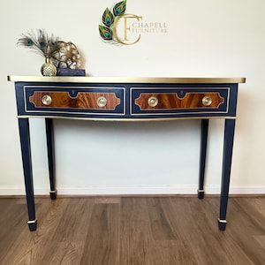 May include: A navy blue console table with gold trim and two drawers. The drawers have a wood veneer with brass pulls. The table has a curved top and sits on four legs.