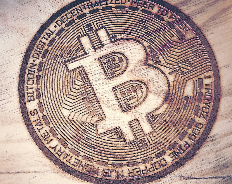 May include: A wooden coin with a Bitcoin logo engraved on it. The coin is inscribed with the words "1 TROYOZ 999 FINE COPPER MUB MONETARY METAL BITCOIN DIGITAL-DECENTRALIZED-PEER TO PEER".