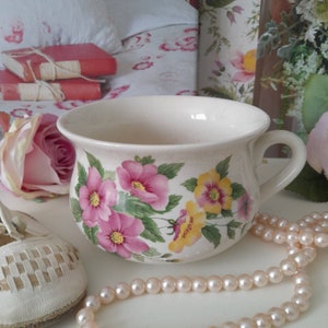 Könnte beinhalten: Eine weiße Keramiktasse mit einem Blumendesign mit rosa und gelben Rosen. Die Tasse hat einen Henkel und steht auf einer weißen Oberfläche mit einer Perlenkette.