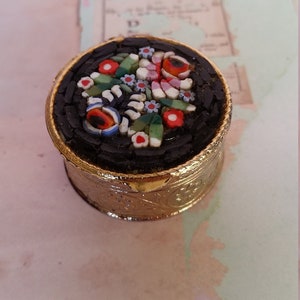 May include: A small, round, gold-colored trinket box with a mosaic floral design on the lid. The mosaic features red, pink, white, blue, and green flowers on a black background.