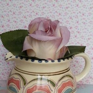 May include: A single, pale purple rose with green leaves sits in a cream-colored ceramic pitcher with a colorful, geometric design.