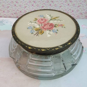 May include: A vintage glass and metal trinket box with a floral design on the lid. The lid is made of a cream-colored fabric with a floral embroidery pattern in pink, blue, and yellow. The box has a clear glass base with a decorative, fluted design.