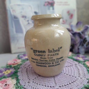 May include: A small, ceramic jar with a white glaze and blue lettering. The jar is labeled "green label" curry paste, made from genuine Madras curry powder. The jar is made by L.A. Sharwood & Co. Ltd., London, England.